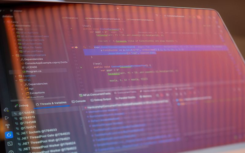 A laptop screen photographed at an angle, showing a code editor with C# unit tests, a project tree listing dependencies and a LICENSE file, and a debugger panel of .NET thread pool workers.