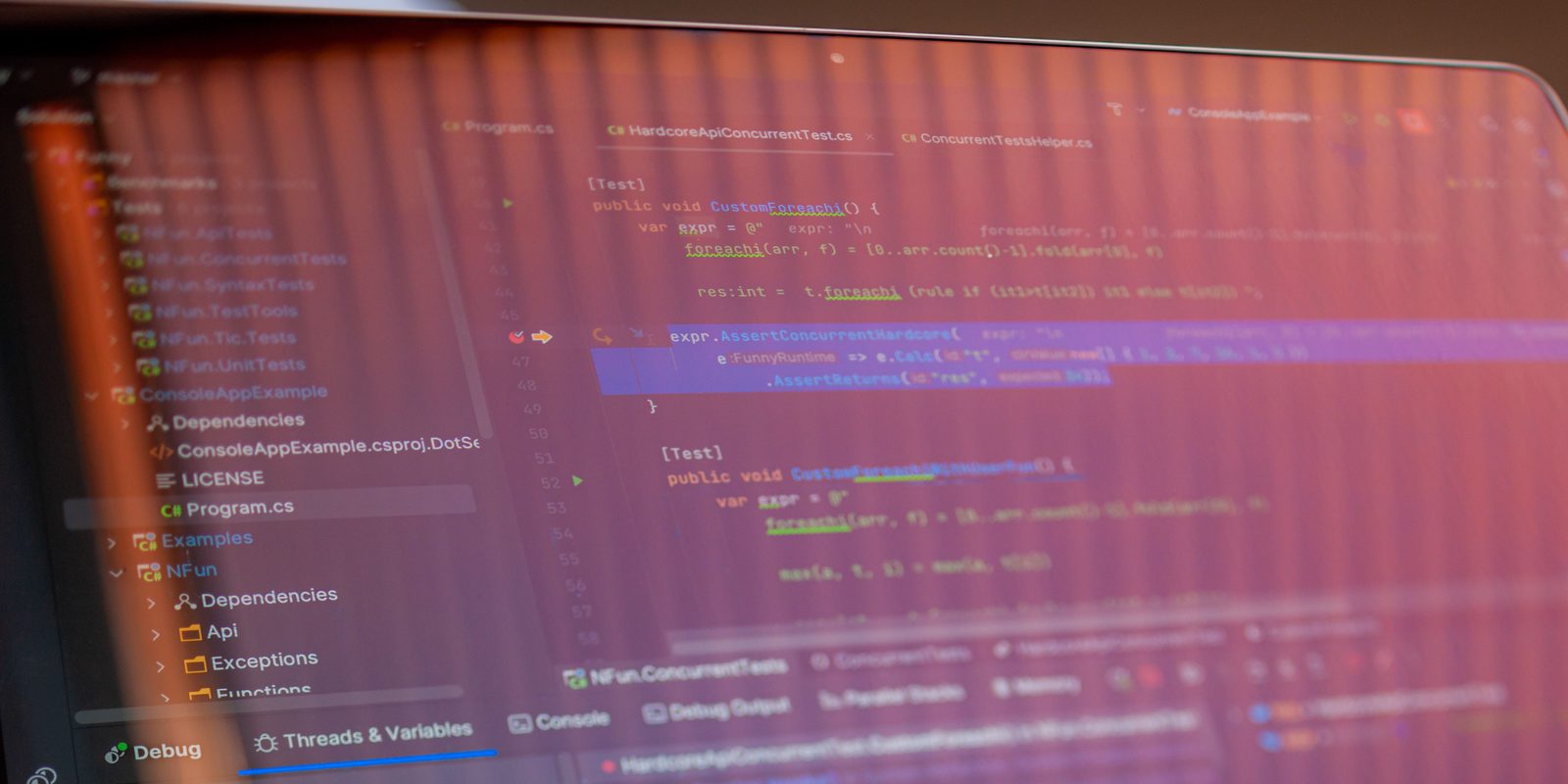 A laptop screen photographed at an angle, showing a code editor with C# unit tests, a project tree listing dependencies and a LICENSE file, and a debugger panel of .NET thread pool workers.